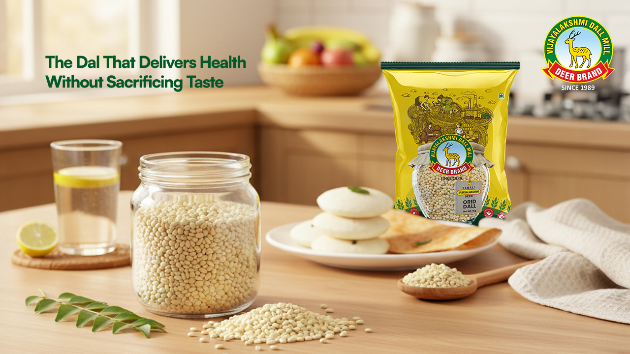 Vijayalakshmi Deer Brand Urad Dal pack displayed on a warm kitchen countertop with a glass jar of whole urad dal, idlis and dosa on a plate, a spoonful of dal, curry leaves, and a glass of lemon water, conveying healthy South Indian breakfast ingredients and natural freshness.