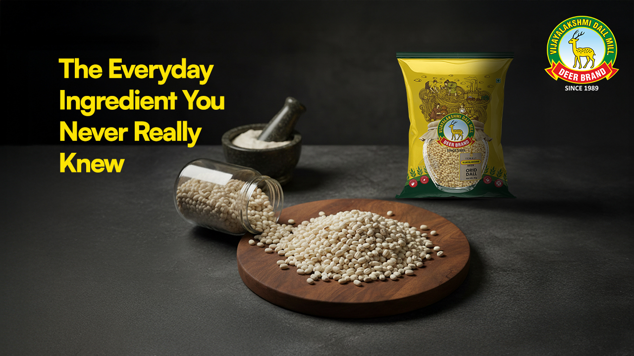A premium Vijayalakshmi Deer Brand Urid Dal pack placed on a dark kitchen surface, with a wooden board full of spilled urad dal beside a glass jar and a stone mortar. Yellow headline reads ‘The Everyday Ingredient You Never Really Knew,’ highlighting the purity and traditional quality of the product.