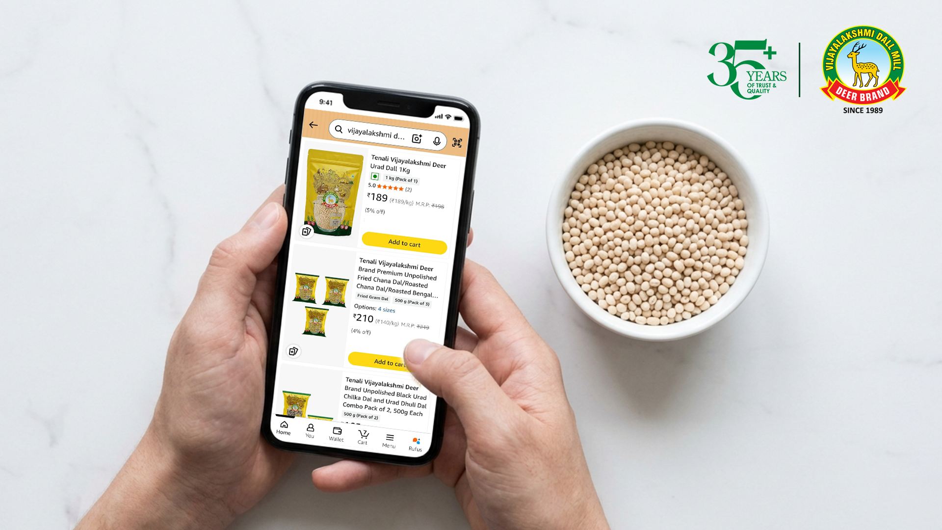 A person holds a smartphone displaying the Vijayalakshmi Deer Brand Urad Dal product listing on an online store. Next to the phone, a white bowl filled with raw urad dal sits on a light-colored surface. The image also includes a 35+ years of trust and quality logo and the Vijayalakshmi Deer Brand logo at the top right.