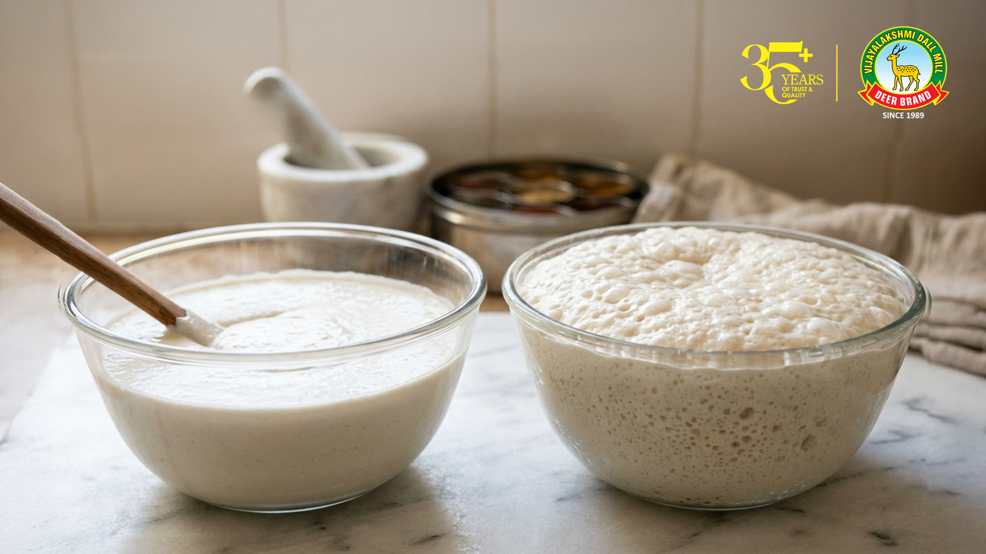 Two glass bowls of idli batter on a kitchen countertop—one with a flat, dense batter that has not fermented well, and the other properly fermented with a light, airy, bubbly texture—highlighting the contrast, with a mortar and spices softly blurred in the background and a Vijayalakshmi Deer Brand logo in the corner.