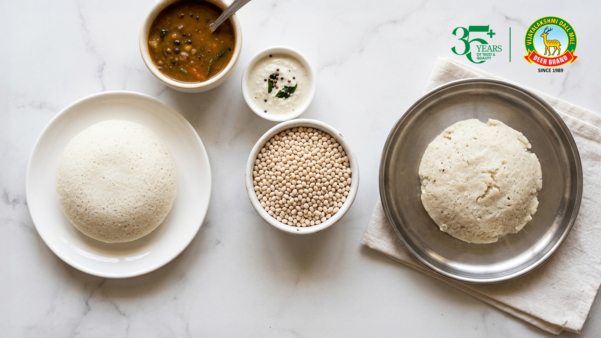 Top-down view of a clean white marble surface featuring South Indian breakfast elements: a soft, spongy idli on a white plate, a bowl of sambar with vegetables, a small bowl of coconut chutney with mustard seeds and curry leaves, and a bowl of whole urad dal. On the right, a metal bowl holds a misshapen idli that hasn’t risen or formed properly, placed on a folded cloth. Brand logos appear in the top-right corner.