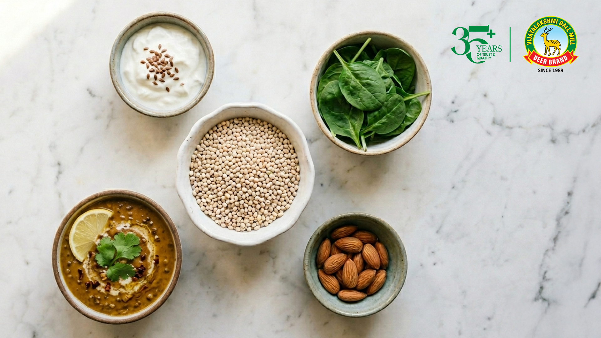 Top-down view of a clean marble surface with five small bowls arranged neatly: a central bowl filled with white lentils (urad dal), surrounded by bowls of creamy yogurt topped with seeds, fresh spinach leaves, whole almonds, and a spiced dal dish garnished with lemon slices and coriander. In the top right corner, a “35+ Years of Trust & Quality” badge appears alongside the Vijayalakshmi Deer Brand logo (Since 1989).