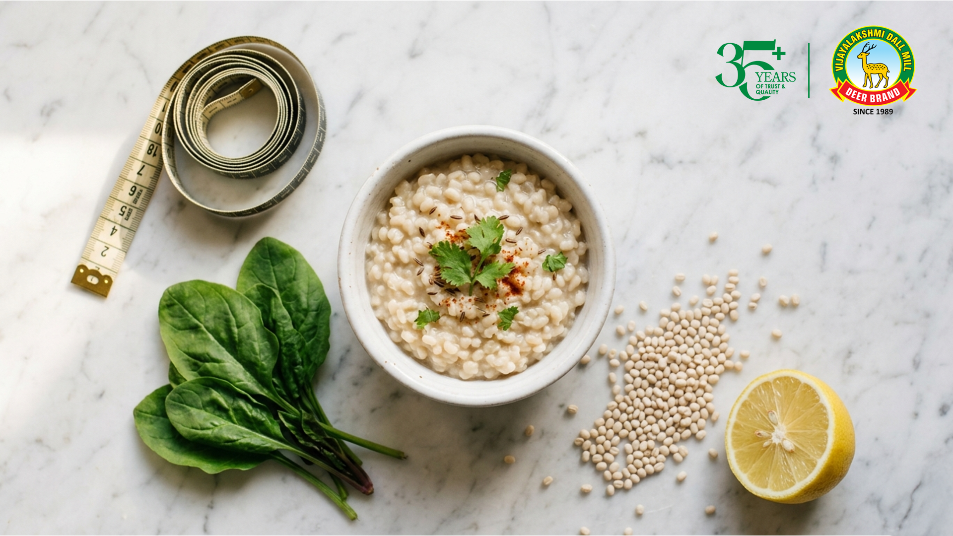 Top-down view of a white bowl of creamy urad dal porridge garnished with coriander and spices, placed on a marble surface with scattered urad dal grains, fresh spinach leaves, a halved lemon, and a measuring tape, alongside Vijayalakshmi Deer Brand logo and “35+ years of trust & quality” mark.